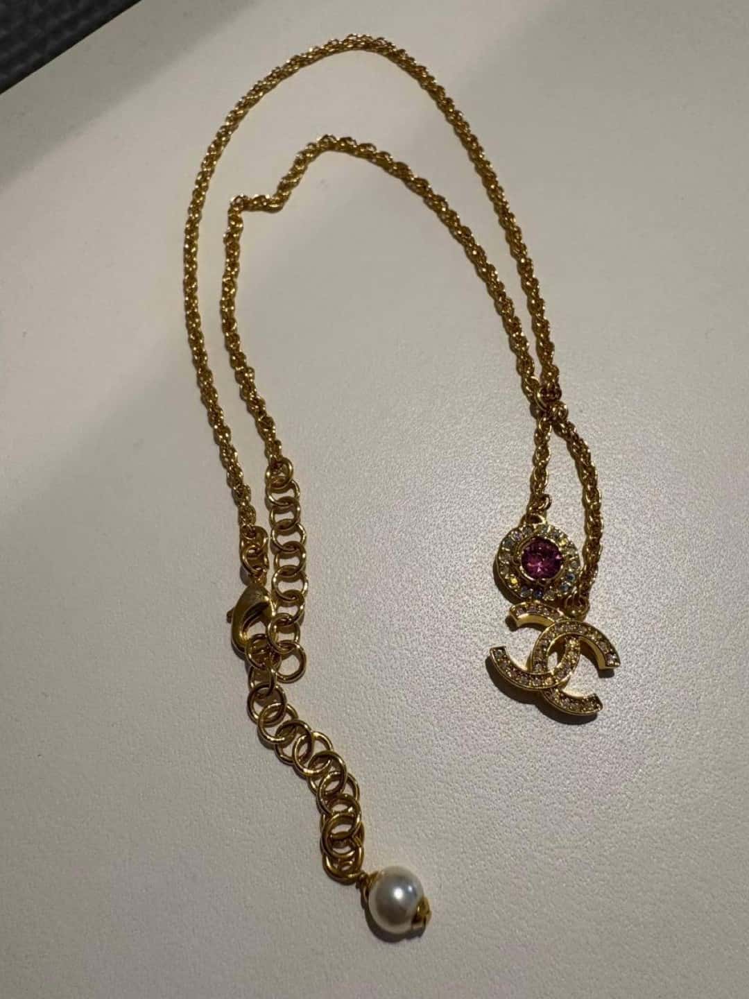 Phone photograph of a gold pendant necklace with a pink stone and pearl-ended extender on a plain surface